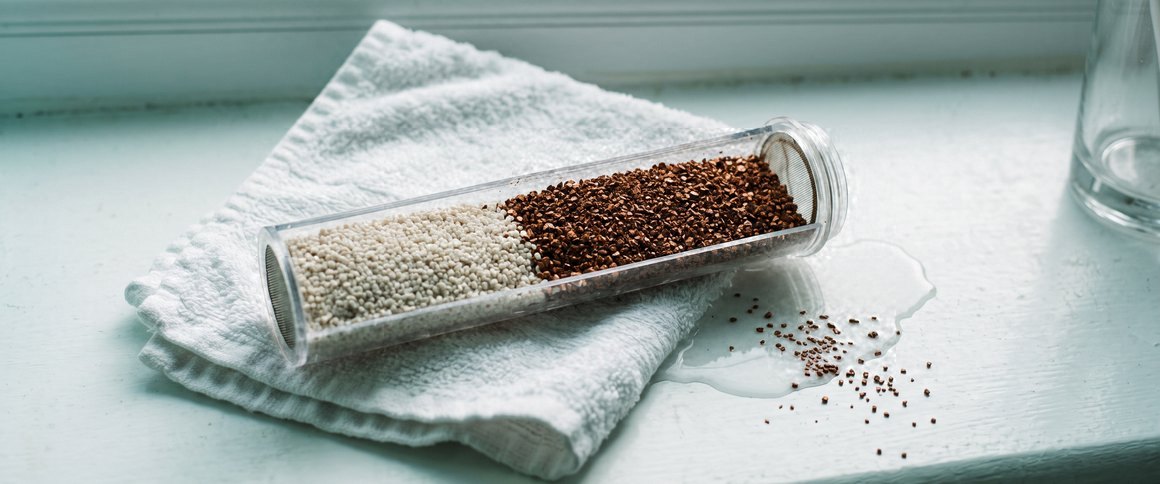 A clear filter cartridge lying open on a folded white towel by a bathroom window, white granules in one half and copper-brown granules in the other, a few spilled on the sill.