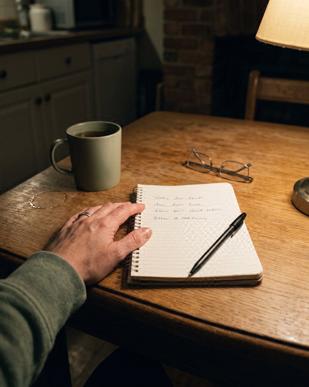 From above at night: a spiral notepad on a kitchen table with a few soft handwritten lines and a pen across it, a hand resting on the page, a mug of tea under lamplight.