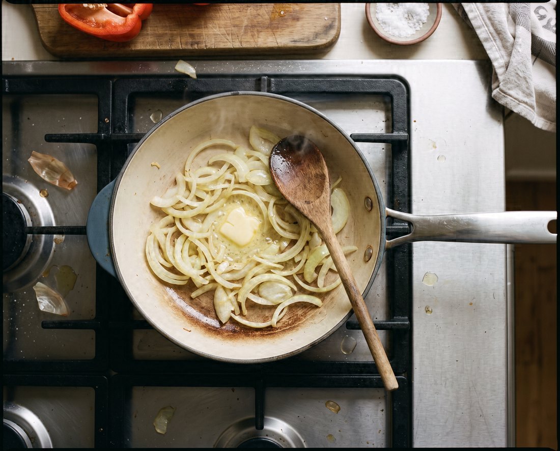 A skillet seen from straight above on a gas hob, onions softening in butter on its cream cooking surface, a wooden spoon resting on the rim.