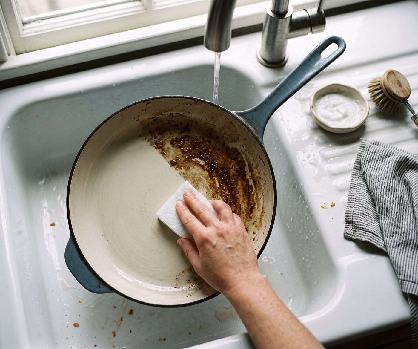 A cream skillet in a white sink from above, the left half scrubbed back to pale cream and the right half still brown with film, a hand working a pad at the line between them, a small dish of white paste on the drainer.